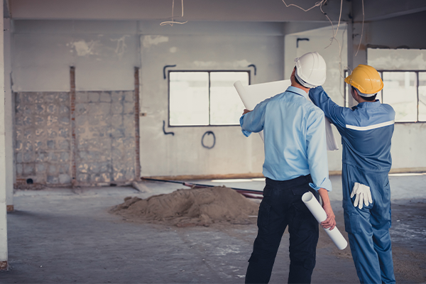 Two construction workers in hard hats review blueprints inside a partially renovated room with exposed wiring and building materials.